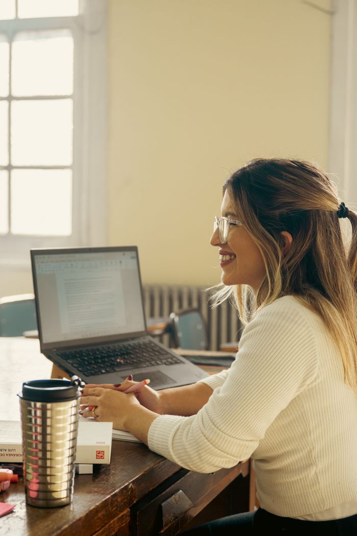 Smiling female teacher at desk with laptop in classroom setting during the day.